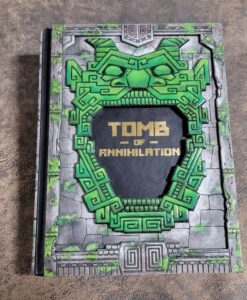 Dungeons and Dragons Tomb of Annihilation, wrapped in black leather, framed in wood etched like stone with a vibrant green face -- mouth opened like a cave.