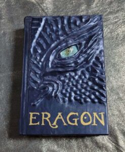 A blue leather bound Eragon by Christopher Paolini. The cover is ridged with the face of a dragon and a piercing blue eye.