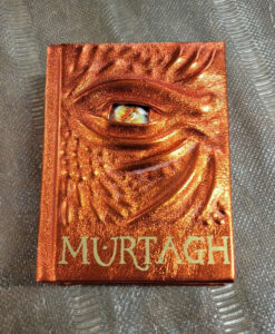 A shiny orange leather bound Murtagh, by Christopher Paolini. The cover is raised with a ridged dragon face and a luminous red and yellow eye.